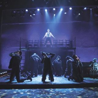 An a scene from TCU's presentation of the musical Spring Awakening, an ensemble of actors in period costume kneel while a male soloist sings under a blue spotlight on an elevated platform.